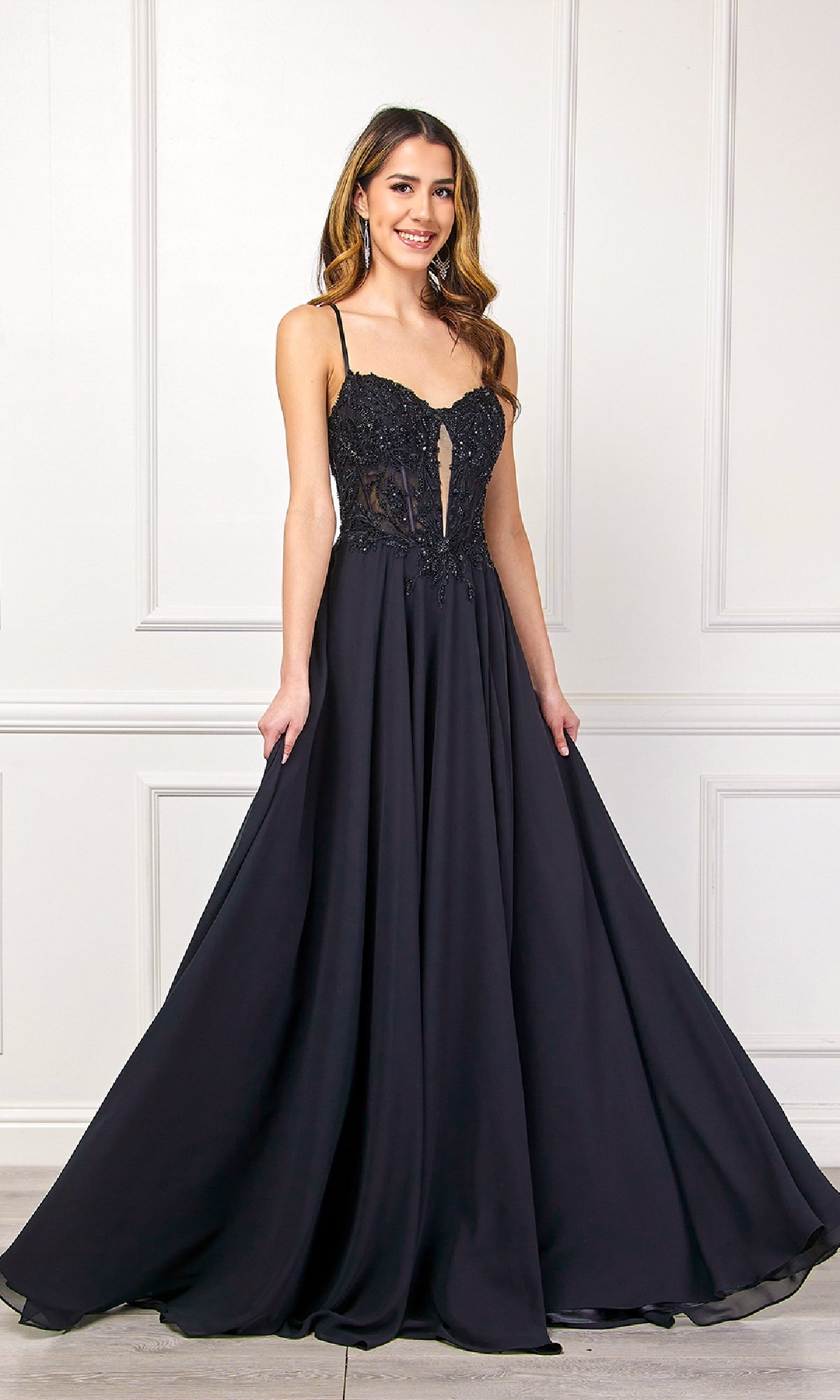 A woman smiles in front of a white paneled wall, wearing the Amelia Couture 7066 Long Black Corset Prom Dress with a fitted bodice and flowing skirt.